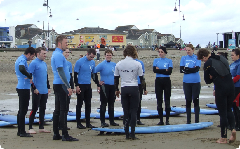 Instructor briefing a group of adult surfers standing beside boards on the beach before a lesson.