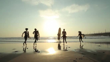Silhouetted group of surfers running along the beach toward the ocean at sunset carrying boards.