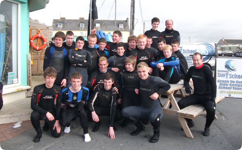 Group of teenage surfers in wetsuits posing together outside a surf school building.
