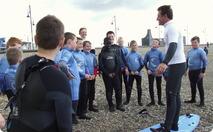 Surf instructor briefing a group of teens in wetsuits on a pebble beach before a lesson.