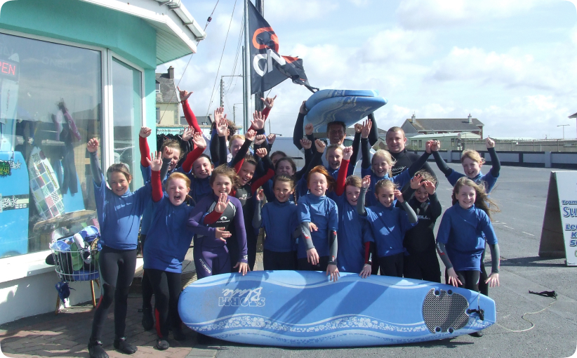 Group of children in wetsuits celebrating outside a surf school with a surfboard raised in front.