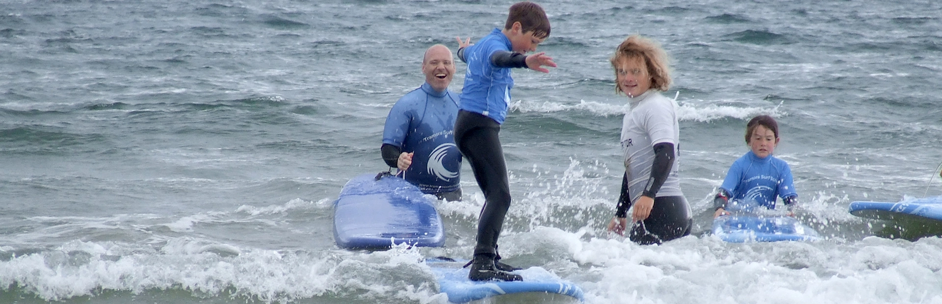 Child riding a small wave on a surfboard while instructors and other children watch nearby.