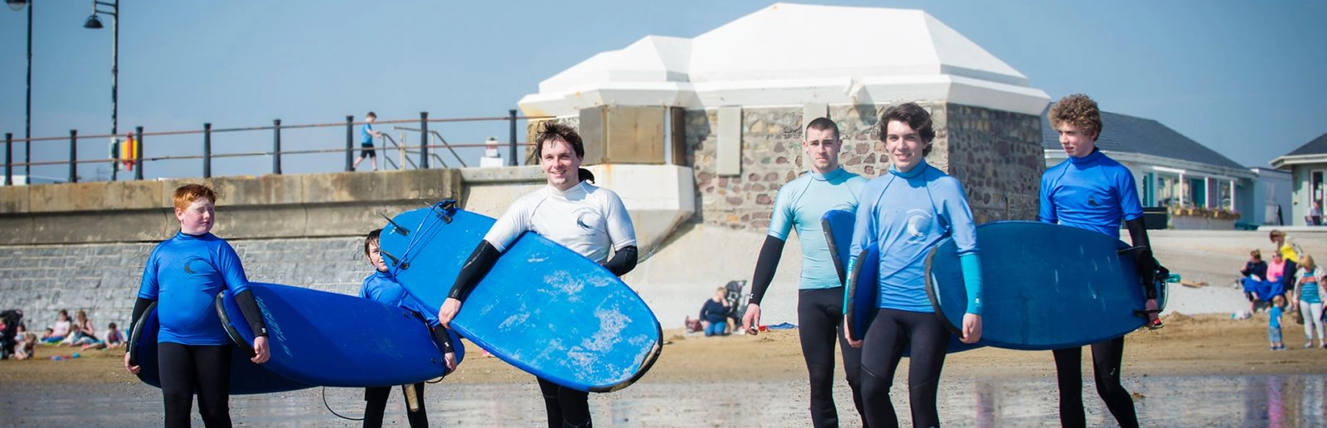 Group of beginner surfers in wetsuits carrying boards while walking along the beach.