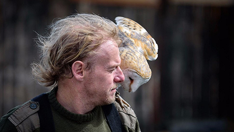 Man with an owl perched on his shoulder outdoors.