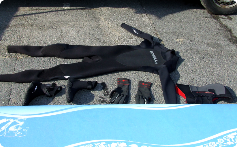 Surf hire package laid out on ground with wetsuit, gloves, boots, hood, and soft surfboard.