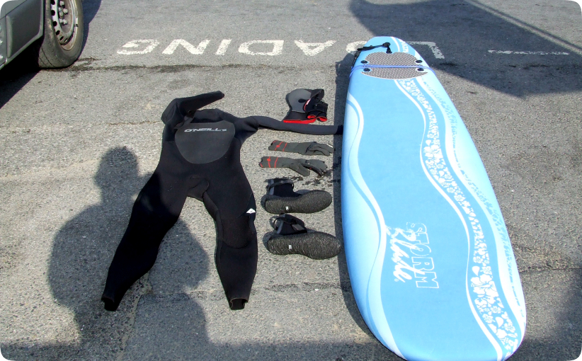 Surf hire package laid out on ground with wetsuit, gloves, boots, hood, and soft surfboard.