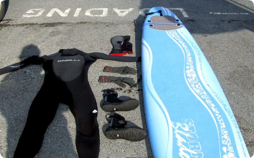 Surf hire package laid out on ground with wetsuit, gloves, boots, hood, and soft surfboard.