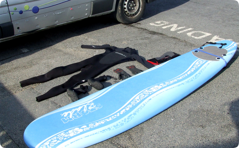 Surf hire package laid out on ground with wetsuit, gloves, boots, hood, and soft surfboard.