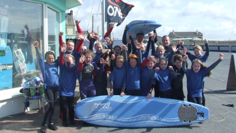 Group of happy children in wetsuits celebrating outside a surf shop with surfboards.