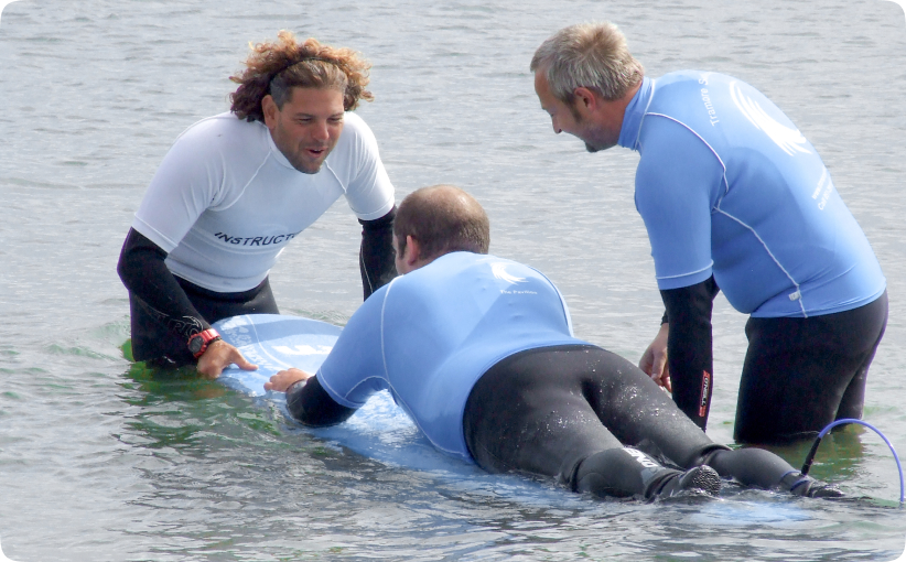 Surf instructors assisting an adult learner lying on a board in shallow water during a private lesson.