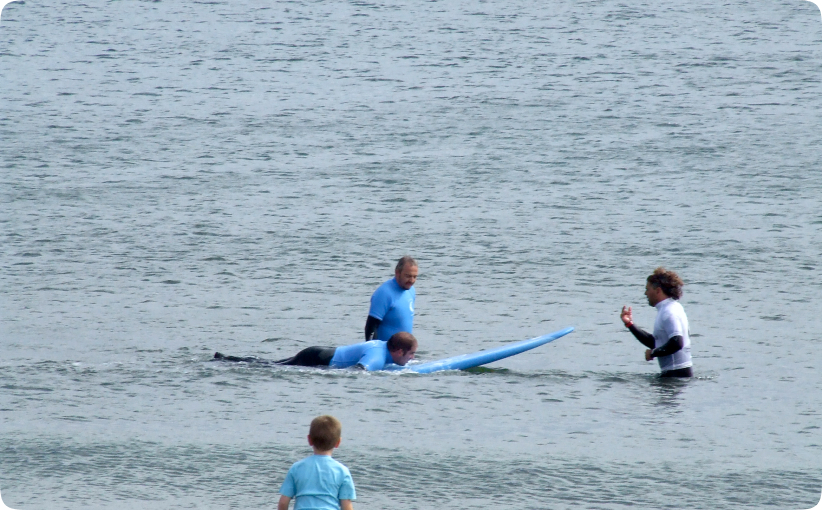 Surf instructor giving guidance to a student lying on a surfboard in shallow water during a lesson.