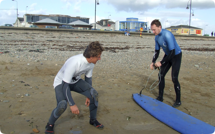 Surf instructor helping a student attach a leash to a board on the beach before a lesson.