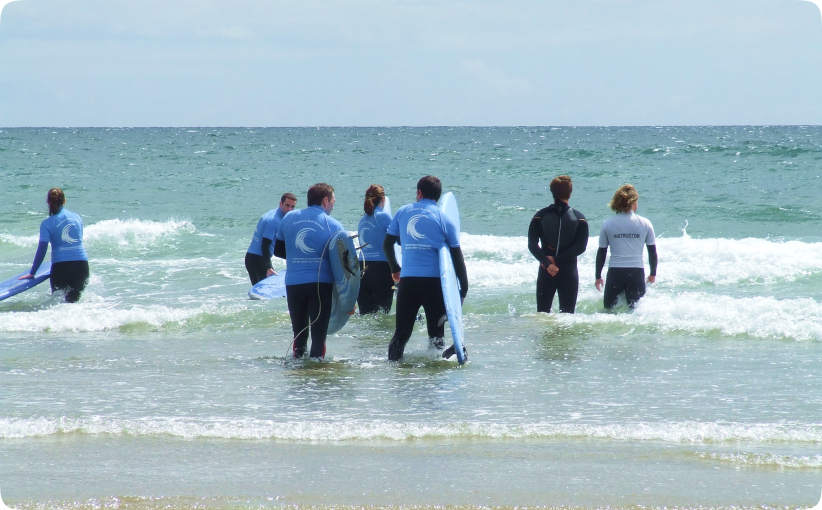 Group of beginner surfers wading into the ocean with boards alongside an instructor during a lesson.