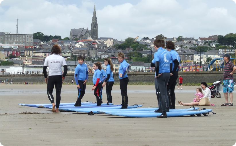 Instructor briefing a group of young surfers standing beside boards on the beach before entering the water.