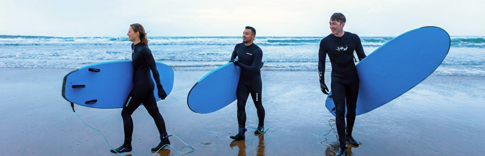 Three surfers in wetsuits carrying boards while walking along the shoreline after surfing.
