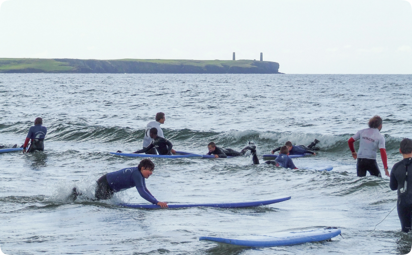 Group of students in wetsuits practising surfing in small waves while instructors supervise in the water.
