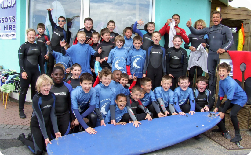 Large group of schoolchildren in wetsuits cheering and smiling while gathered around a surfboard outside a surf school.