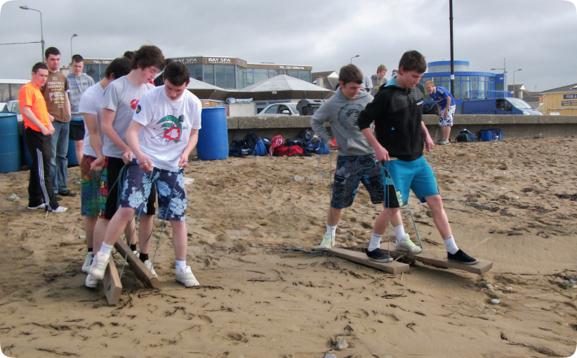 Group of teenage boys balancing on wooden team-building skis during a beach activity.