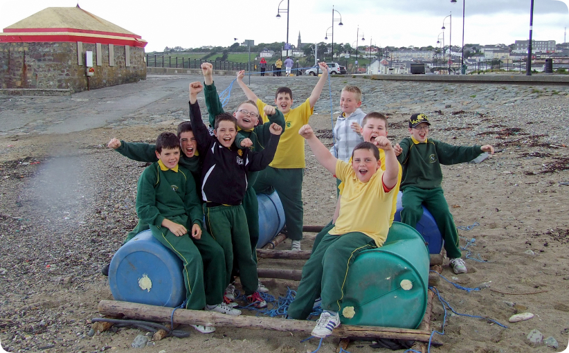 Group of schoolchildren sitting on barrel rafts on the beach, raising their arms and cheering during a team-building activity.