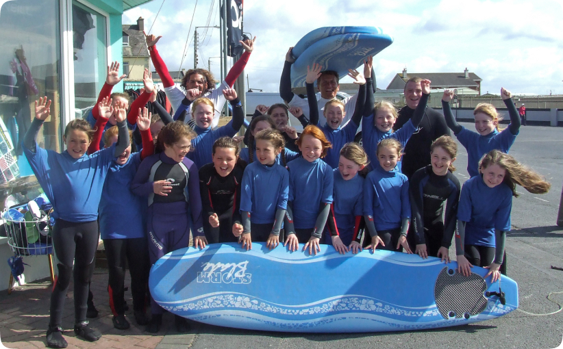 Group of schoolchildren in blue wetsuits smiling and raising their hands while holding a surfboard outside a surf school.
