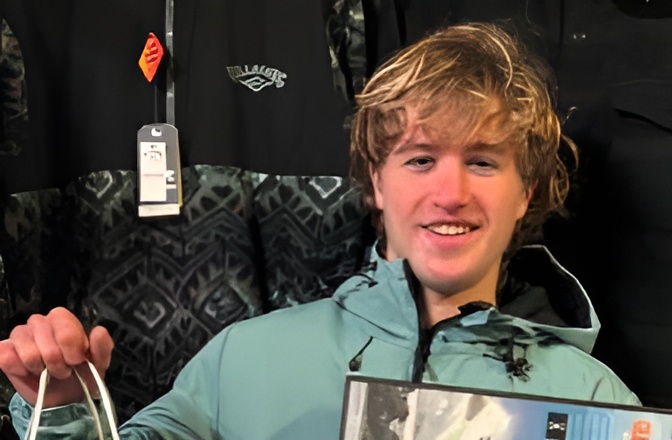 Portrait of a young surf shop staff member holding shopping bags in front of wetsuits.