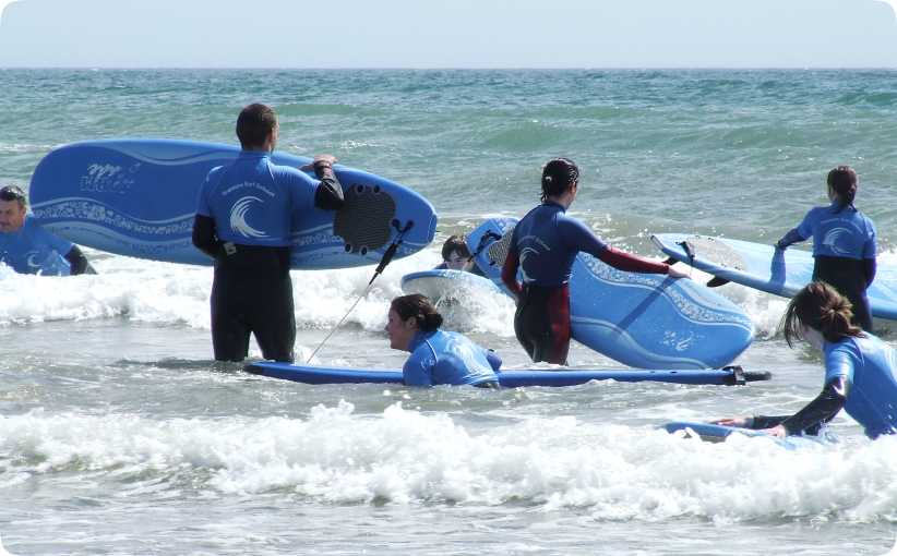 Group of beginner surfers in wetsuits holding softboards in shallow water during a lesson.