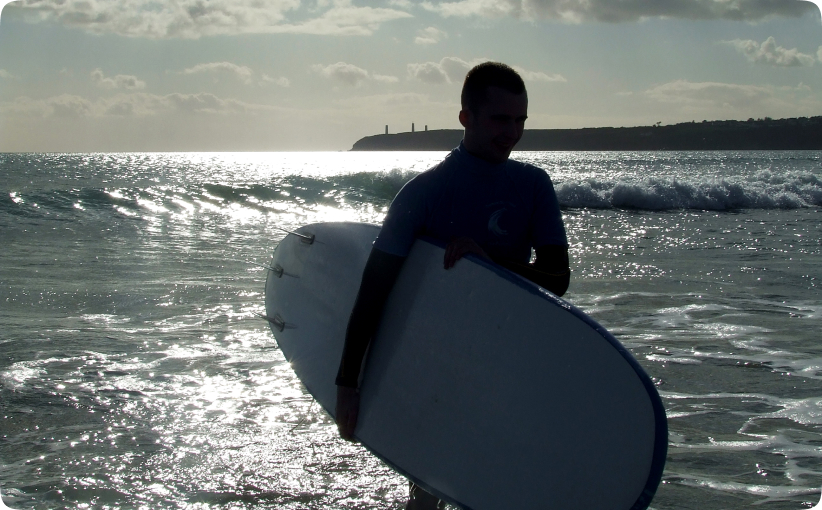 Surfer carrying a softboard in shallow water with sunlight reflecting on the sea.