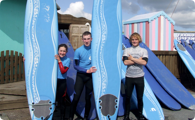 Three young surfers standing between upright softboards outside a surf shop.