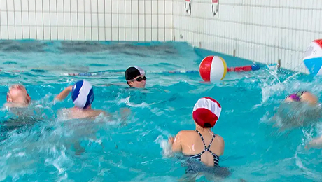 Children wearing swim caps playing with a beach ball in an indoor swimming pool.