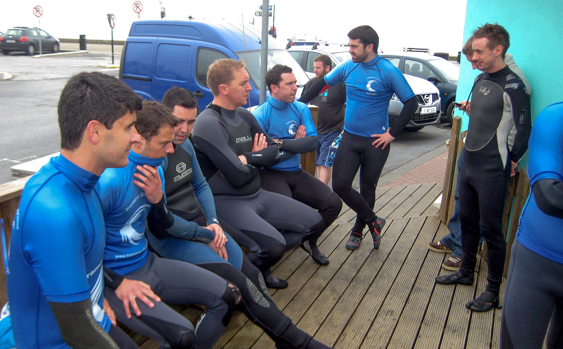 A stag party group in wetsuits gathered outside a surf school, listening to an instructor give a briefing before heading into the water.