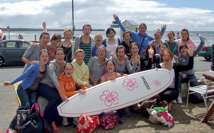 Group celebrating a hen party on the beach, posing with a surfboard and cheering.