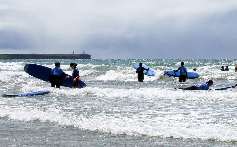 A hen party group in matching surf tops wading into the sea with their boards, laughing and supporting each other as they begin their surf lesson, with waves breaking and the coastline in the background.