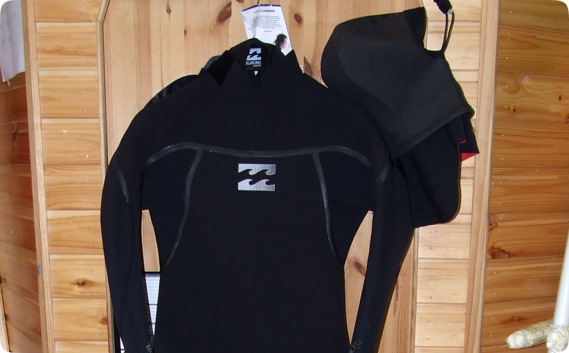 Black full-body wetsuit with hood hanging on a wooden wall inside a surf shop.