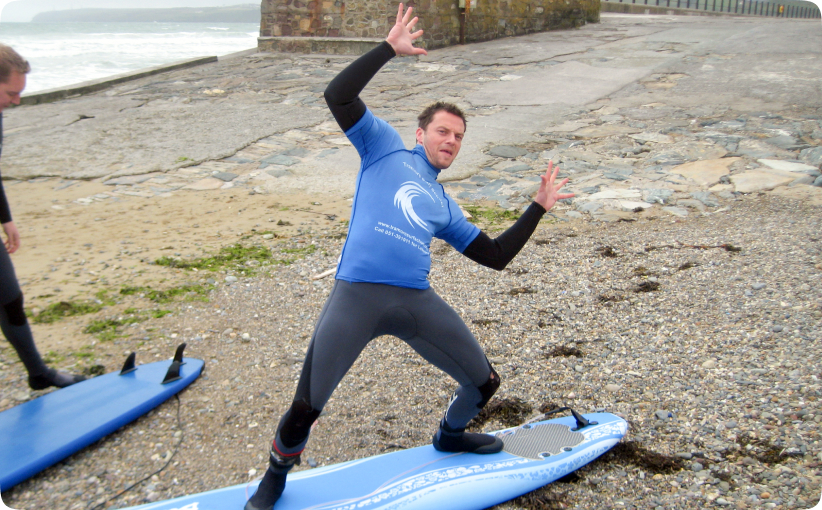 Man in a wetsuit demonstrating a surfing stance on a board on the beach.