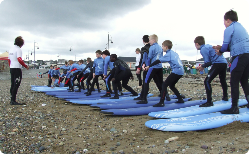 Group of children in blue wetsuits practising standing on surfboards on the beach while an instructor guides them.