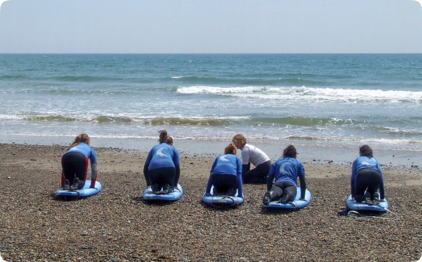 Group of children in blue wetsuits kneeling on surfboards at the shoreline while an instructor guides them before entering the water.