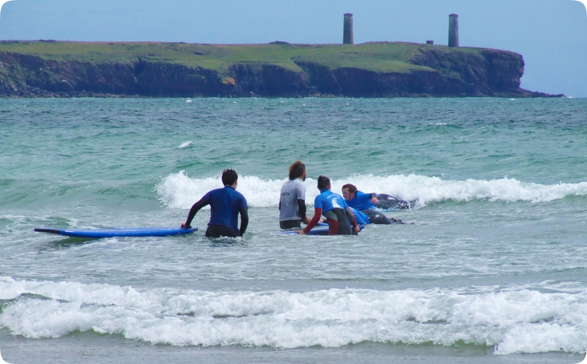 Surf instructor assisting students on surfboards in shallow waves with a rocky coastline in the background.