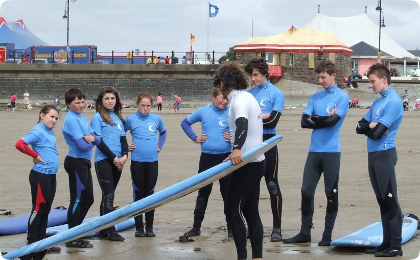 Surf instructor holding a surfboard while speaking to a group of teenagers in blue wetsuits on the beach during a lesson.