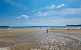 Sandy beach in Tramore with shallow water and a few people walking along the shoreline under a partly cloudy sky.